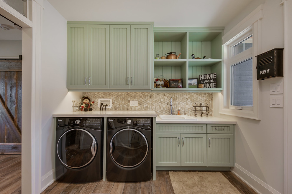 Klassisk inredning av en vita linjär vitt tvättstuga enbart för tvätt, med en nedsänkt diskho, luckor med infälld panel, gröna skåp, mörkt trägolv, en tvättmaskin och torktumlare bredvid varandra och brunt golv