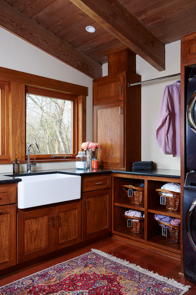 Große Klassische Waschküche in L-Form mit Landhausspüle, Schrankfronten im Shaker-Stil, Granit-Arbeitsplatte, weißer Wandfarbe, Waschmaschine und Trockner gestapelt, hellbraunen Holzschränken und braunem Holzboden in Minneapolis