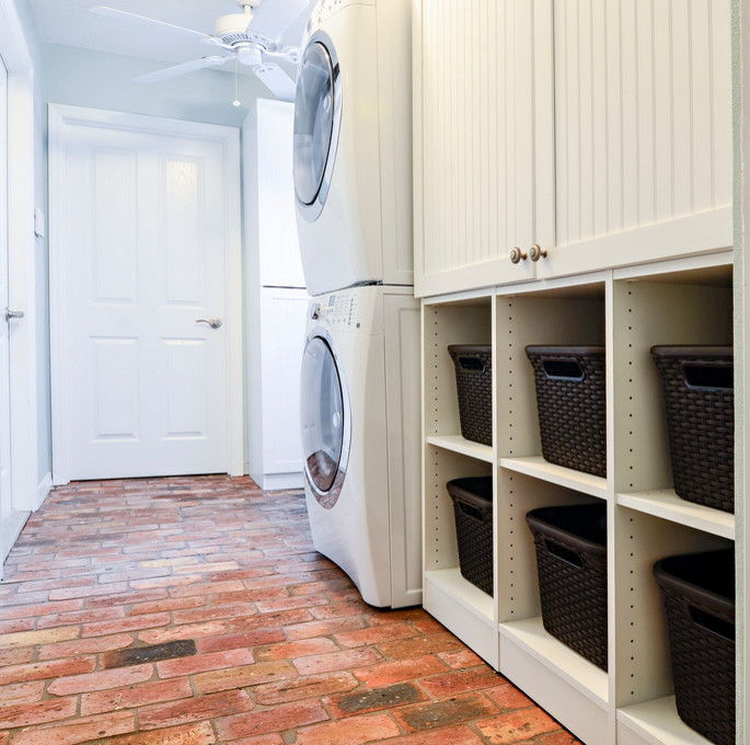 Immagine di una lavanderia multiuso di medie dimensioni con ante bianche, pareti bianche, pavimento in mattoni e lavatrice e asciugatrice a colonna