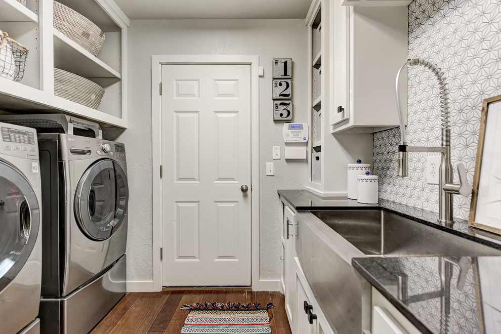 Design ideas for a medium sized traditional galley separated utility room in Oklahoma City with a belfast sink, shaker cabinets, white cabinets, granite worktops, grey walls, medium hardwood flooring, a side by side washer and dryer, brown floors and black worktops.