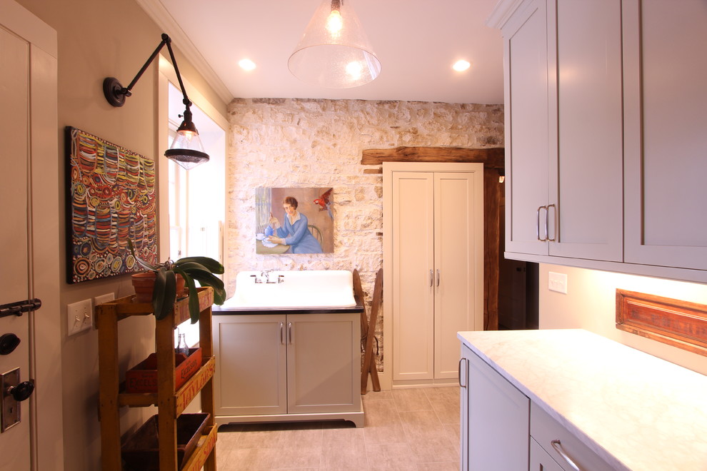 Photo of a medium sized rural galley utility room in Other with a belfast sink, recessed-panel cabinets, granite worktops, beige walls, porcelain flooring, a stacked washer and dryer, brown floors, green worktops and grey cabinets.