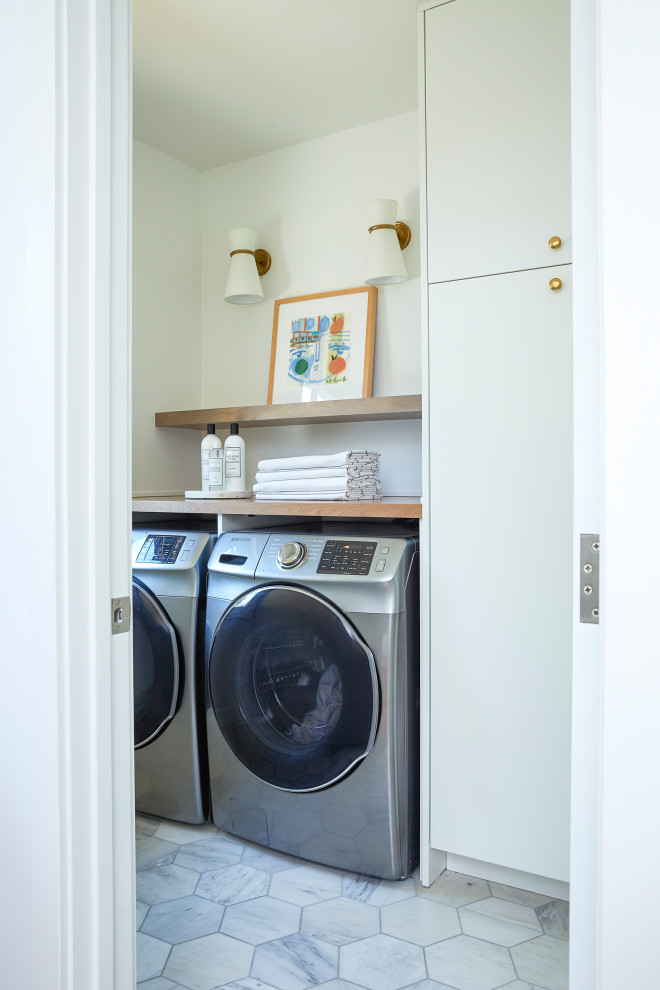 Modern inredning av en liten bruna linjär brunt tvättstuga enbart för tvätt, med släta luckor, vita skåp, träbänkskiva, vita väggar, marmorgolv, en tvättmaskin och torktumlare bredvid varandra och grått golv