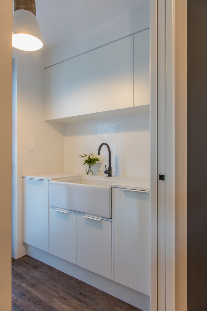 Photo of a small contemporary galley separated utility room in Other with a belfast sink, flat-panel cabinets, white cabinets, engineered stone countertops, white walls, medium hardwood flooring, a stacked washer and dryer, brown floors and white worktops.