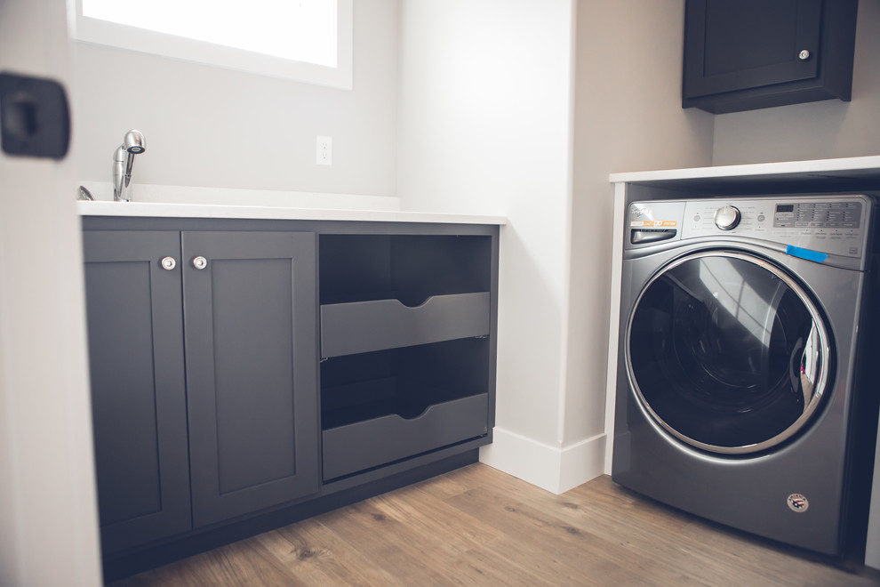 Idéer för mellanstora funkis l-formade tvättstugor enbart för tvätt, med skåp i shakerstil, svarta skåp, grå väggar, mellanmörkt trägolv, en tvättmaskin och torktumlare bredvid varandra och brunt golv