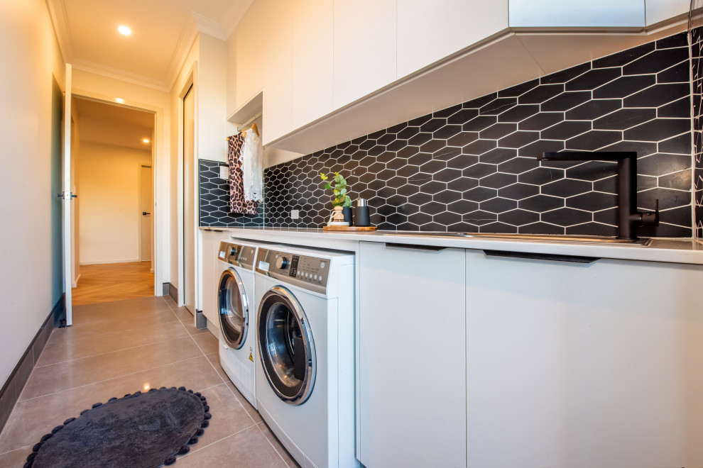 Example of a mid-sized trendy single-wall ceramic tile and gray floor dedicated laundry room design in Other with a single-bowl sink, flat-panel cabinets, white cabinets, granite countertops, black backsplash, mosaic tile backsplash, white walls, a side-by-side washer/dryer and white countertops