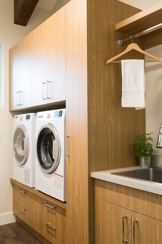 Moderne Waschküche mit flächenbündigen Schrankfronten, hellbraunen Holzschränken, Laminat-Arbeitsplatte, weißer Wandfarbe, dunklem Holzboden, Waschmaschine und Trockner nebeneinander und braunem Boden in Vancouver