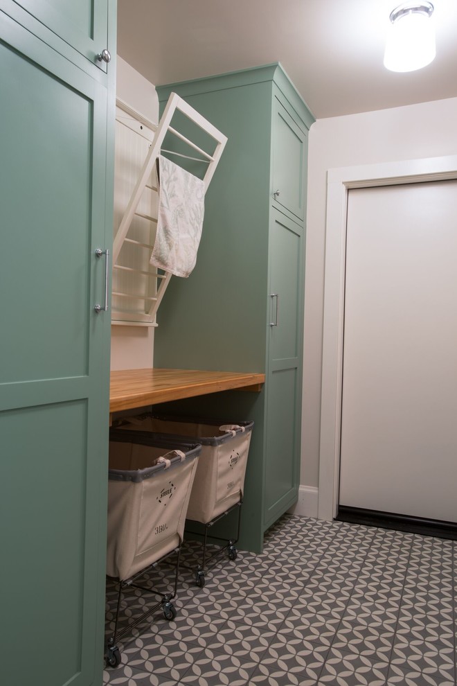 Lantlig inredning av en mellanstor bruna linjär brunt tvättstuga enbart för tvätt, med luckor med infälld panel, gröna skåp, träbänkskiva, vita väggar, klinkergolv i keramik och flerfärgat golv