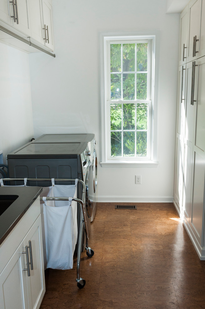 Inredning av en tvättstuga, med luckor med infälld panel, vita skåp, vita väggar, korkgolv, en tvättmaskin och torktumlare bredvid varandra och brunt golv