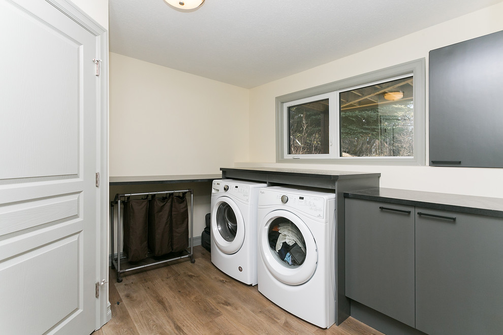 Immagine di una sala lavanderia minimal di medie dimensioni con ante in stile shaker, ante grigie, pareti beige, pavimento in legno massello medio, lavatrice e asciugatrice affiancate e pavimento marrone