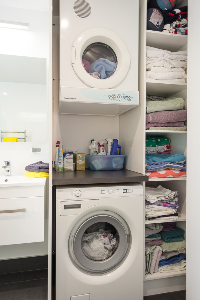 Photo of a small contemporary single-wall utility room in Auckland with flat-panel cabinets, white cabinets, laminate countertops, white walls and a stacked washer and dryer.