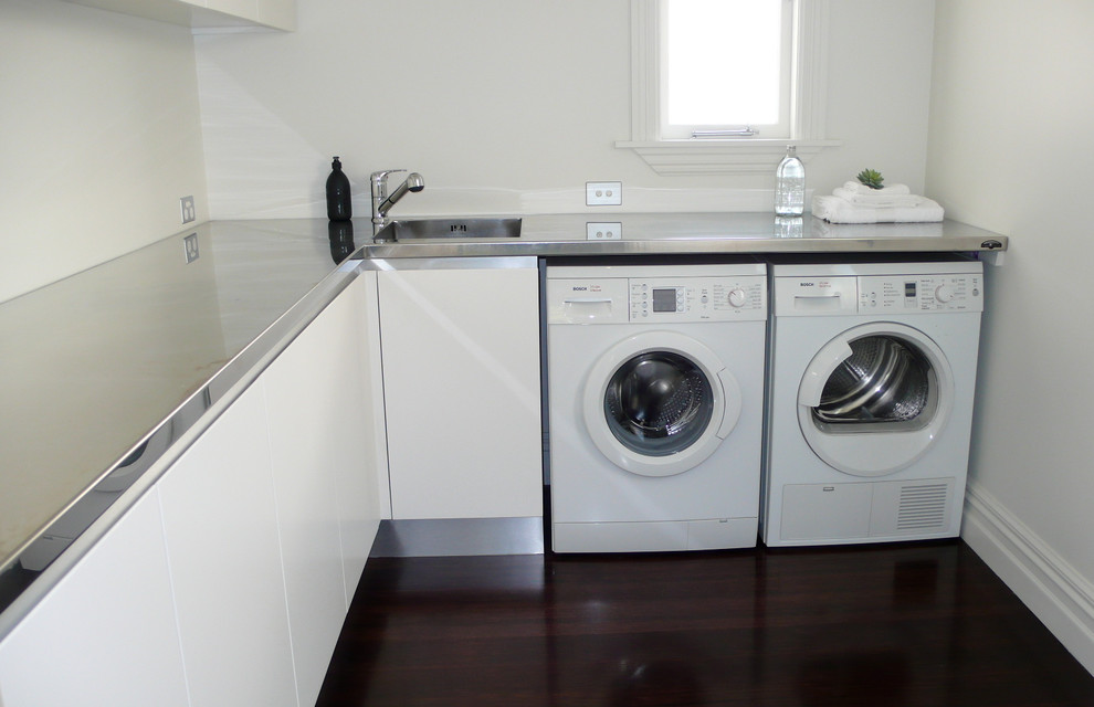 Foto på en funkis l-formad tvättstuga, med en integrerad diskho, släta luckor, vita skåp, bänkskiva i rostfritt stål, vita väggar, mörkt trägolv och en tvättmaskin och torktumlare bredvid varandra