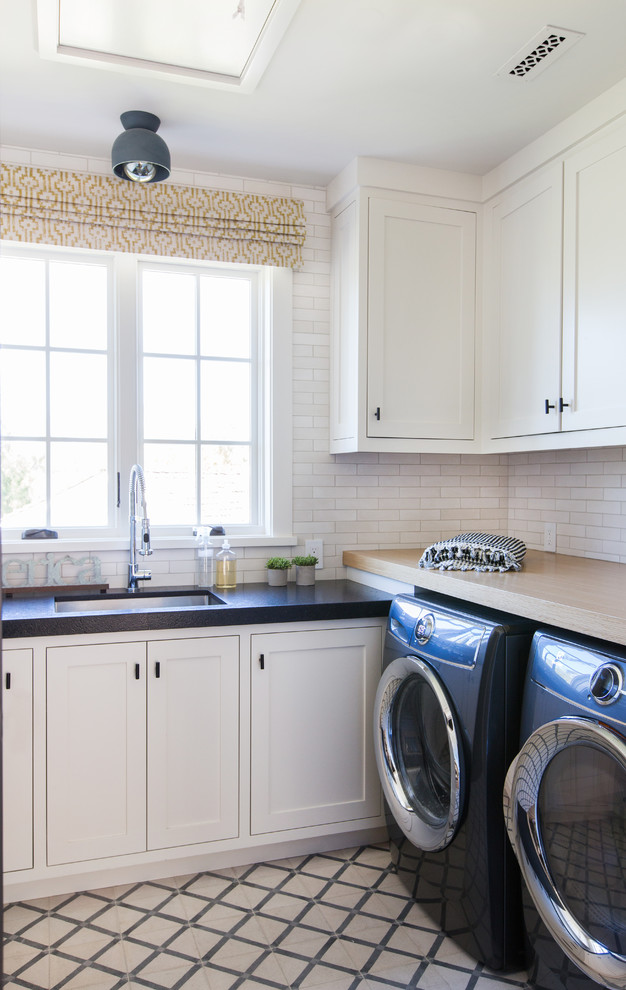Inspiration för maritima l-formade svart tvättstugor enbart för tvätt, med en undermonterad diskho, vita skåp, vita väggar, en tvättmaskin och torktumlare bredvid varandra, beiget golv och skåp i shakerstil