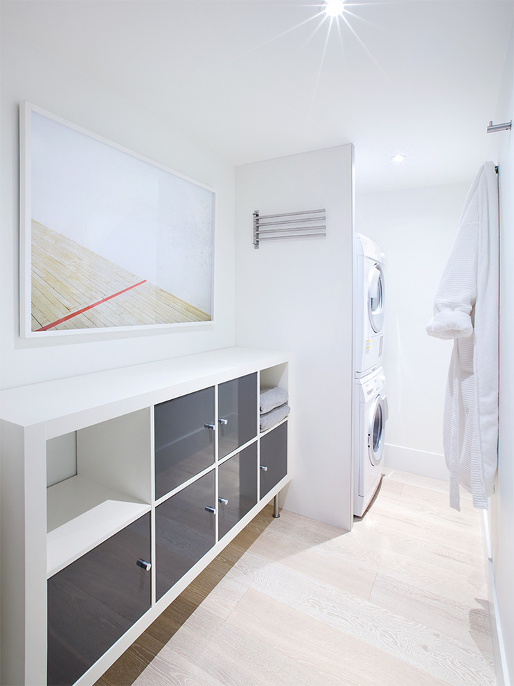 Ejemplo de lavadero lineal contemporáneo con armarios con paneles lisos, puertas de armario grises, paredes blancas, suelo de madera clara y lavadora y secadora apiladas