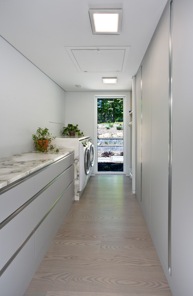 Exempel på en mellanstor modern vita parallell vitt tvättstuga enbart för tvätt, med släta luckor, grå skåp, granitbänkskiva, grå väggar, ljust trägolv, en tvättmaskin och torktumlare bredvid varandra och beiget golv
