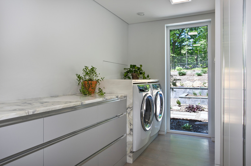 Bild på en mellanstor funkis vita parallell vitt tvättstuga enbart för tvätt, med släta luckor, grå skåp, granitbänkskiva, grå väggar, ljust trägolv, en tvättmaskin och torktumlare bredvid varandra och beiget golv