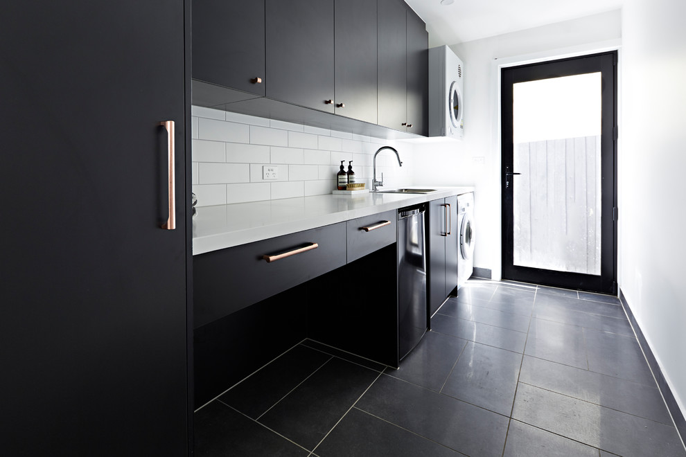 Photo of a medium sized modern single-wall separated utility room in Melbourne with a submerged sink, black cabinets, a stacked washer and dryer, black floors, white worktops, engineered stone countertops and white walls.