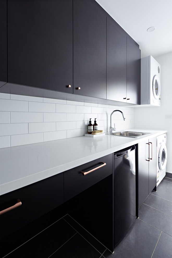 This is an example of a medium sized modern single-wall separated utility room in Melbourne with a submerged sink, black cabinets, engineered stone countertops, white walls, a stacked washer and dryer, black floors and white worktops.