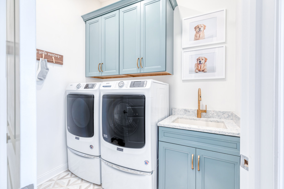 Bild på en vintage grå linjär grått tvättstuga, med en undermonterad diskho, luckor med infälld panel, blå skåp, vita väggar, en tvättmaskin och torktumlare bredvid varandra och vitt golv