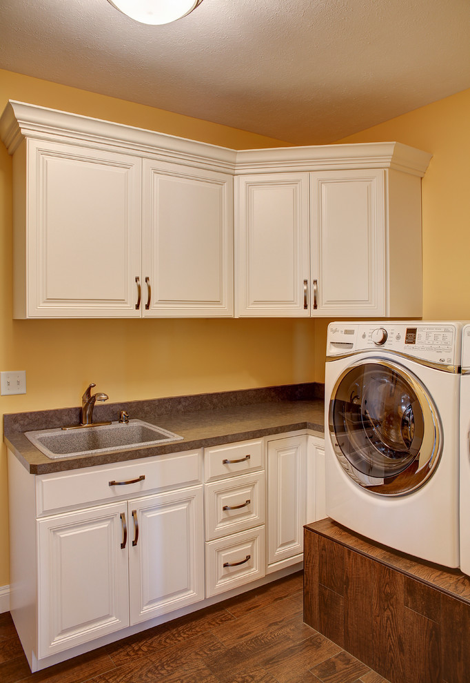 Idéer för en stor klassisk bruna l-formad tvättstuga enbart för tvätt, med en nedsänkt diskho, luckor med upphöjd panel, vita skåp, laminatbänkskiva, gula väggar, en tvättmaskin och torktumlare bredvid varandra, mellanmörkt trägolv och brunt golv