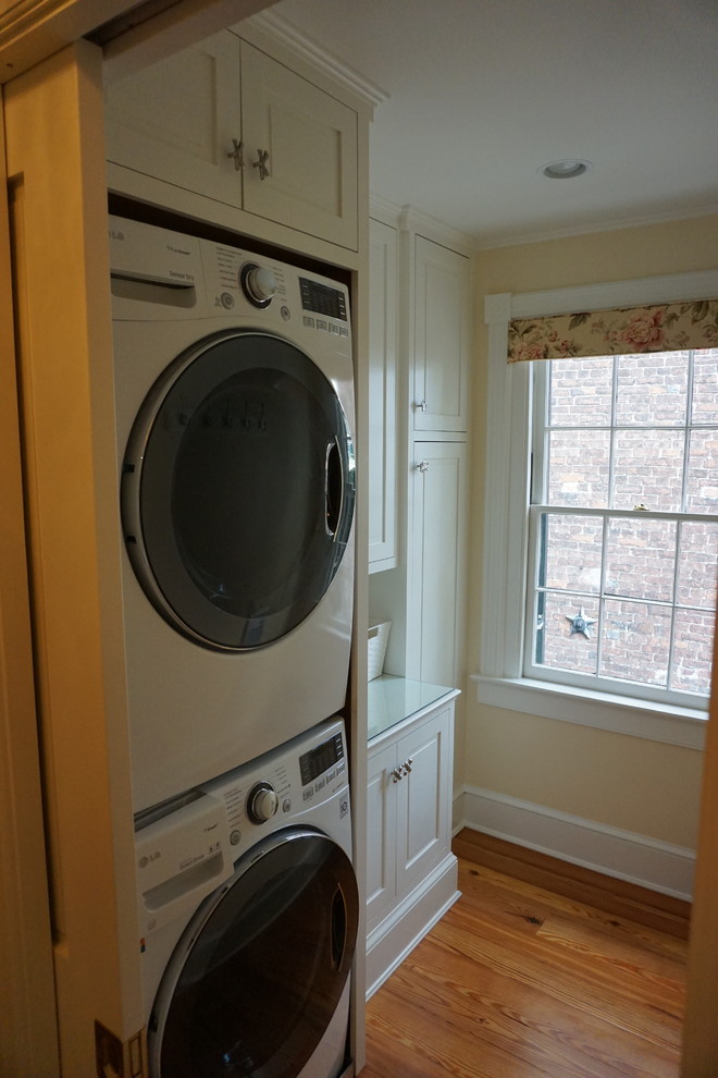 Klassisk inredning av en liten parallell tvättstuga enbart för tvätt, med vita skåp, bänkskiva i glas, beige väggar, mörkt trägolv, en tvättpelare och luckor med upphöjd panel
