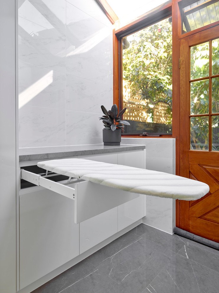 Bild på en mellanstor funkis grå parallell grått tvättstuga enbart för tvätt, med en nedsänkt diskho, släta luckor, vita skåp, bänkskiva i kvarts, vita väggar, klinkergolv i porslin, tvättmaskin och torktumlare byggt in i ett skåp och grått golv
