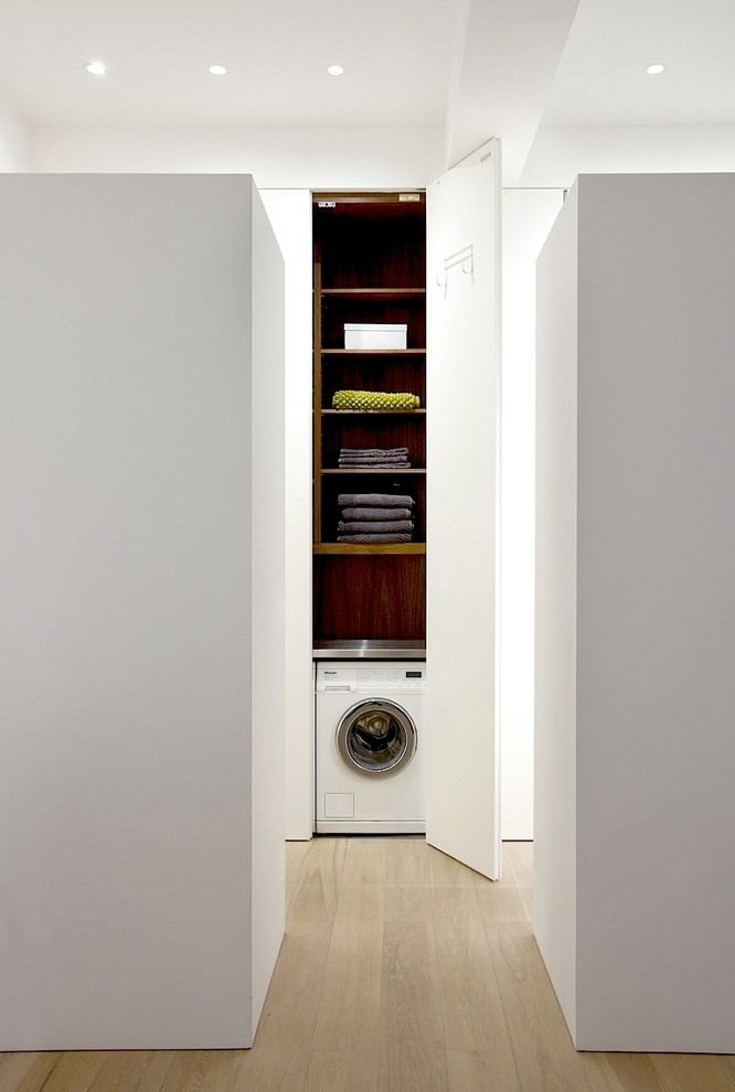 Diseño de armario lavadero moderno pequeño con paredes blancas, suelo de madera clara, armarios con paneles lisos, puertas de armario blancas y lavadora y secadora escondidas