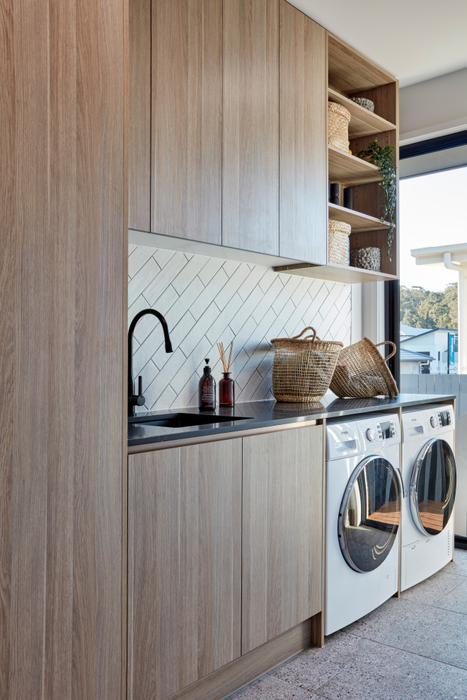 Einzeilige Moderne Waschküche mit Unterbauwaschbecken, flächenbündigen Schrankfronten, hellbraunen Holzschränken, weißer Wandfarbe, Waschmaschine und Trockner nebeneinander, grauem Boden und schwarzer Arbeitsplatte in Melbourne