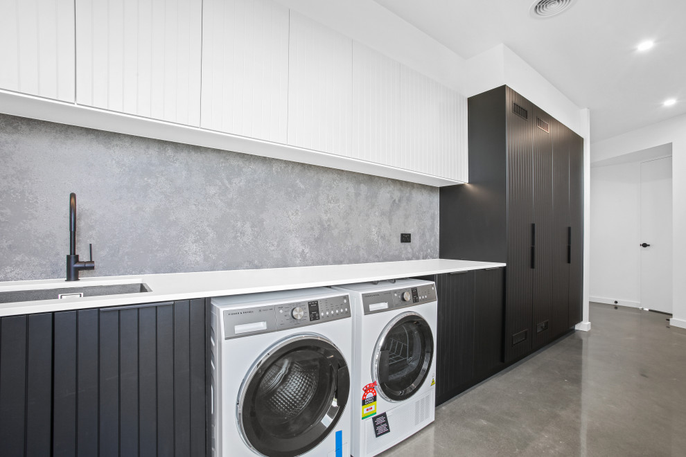 Exempel på en stor modern vita linjär vitt tvättstuga enbart för tvätt, med en undermonterad diskho, luckor med profilerade fronter, svarta skåp, bänkskiva i kvarts, grått stänkskydd, stänkskydd i sten, vita väggar, betonggolv, en tvättmaskin och torktumlare bredvid varandra och grått golv