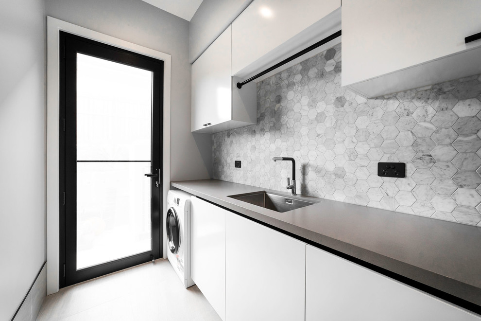 Foto på ett mellanstort funkis vit parallellt grovkök, med en nedsänkt diskho, släta luckor, vita skåp, bänkskiva i betong, grå väggar, betonggolv, en tvättmaskin och torktumlare bredvid varandra och grått golv