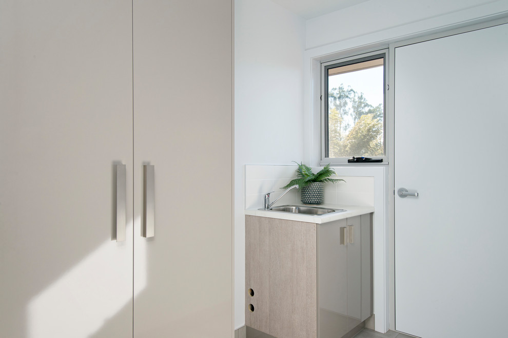 Photo of a small contemporary galley utility room in Other with a built-in sink, recessed-panel cabinets, grey cabinets, laminate countertops, white walls, vinyl flooring, a stacked washer and dryer, brown floors and white worktops.