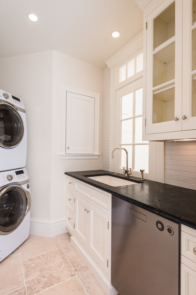 Immagine di una lavanderia multiuso stile marino con lavello sottopiano, ante bianche, pavimento in pietra calcarea, lavatrice e asciugatrice a colonna e pareti beige