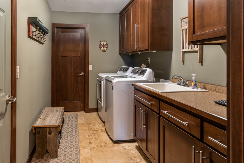 ミネアポリスにある中くらいなカントリー風のおしゃれな家事室 (ll型、ドロップインシンク、シェーカースタイル扉のキャビネット、濃色木目調キャビネット、クオーツストーンカウンター、茶色いキッチンパネル、クオーツストーンのキッチンパネル、緑の壁、トラバーチンの床、左右配置の洗濯機・乾燥機、マルチカラーの床、ベージュのキッチンカウンター) の写真