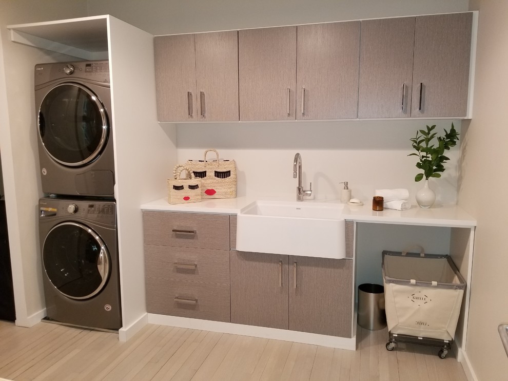 Example of a mid-sized trendy single-wall light wood floor and beige floor dedicated laundry room design in New York with a farmhouse sink, flat-panel cabinets, gray cabinets, quartz countertops, gray walls, a stacked washer/dryer and white countertops