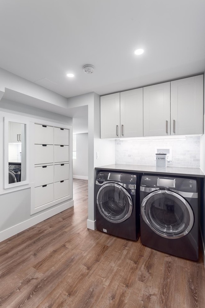 This is an example of a medium sized contemporary single-wall separated utility room in Vancouver with flat-panel cabinets, white cabinets, composite countertops, white walls, medium hardwood flooring, a side by side washer and dryer and brown floors.