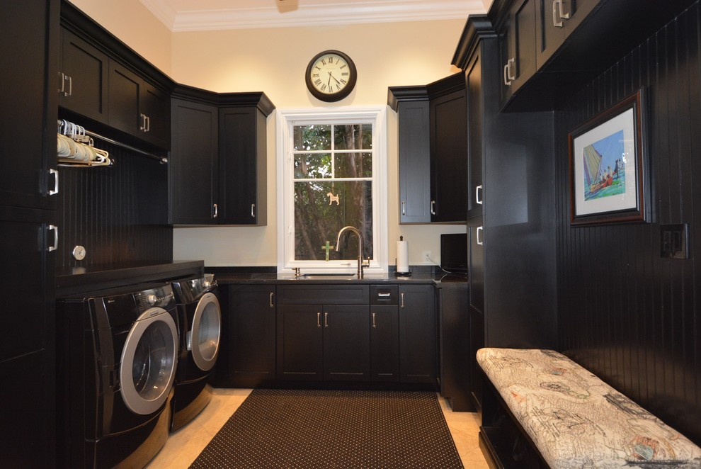 Modelo de lavadero en U clásico renovado con puertas de armario negras, paredes beige, suelo de travertino, lavadora y secadora juntas y suelo beige