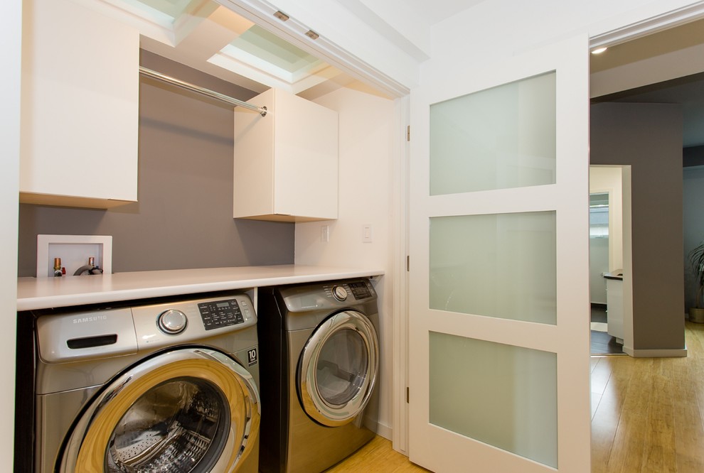Bild på en mellanstor funkis linjär tvättstuga enbart för tvätt, med släta luckor, vita skåp, bänkskiva i kvarts, vita väggar, bambugolv och en tvättmaskin och torktumlare bredvid varandra