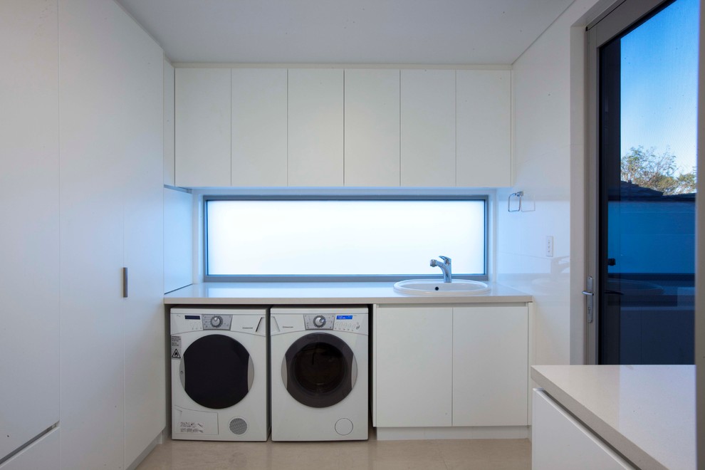 Foto på en mellanstor funkis linjär tvättstuga, med en nedsänkt diskho, släta luckor, vita skåp, vita väggar och en tvättmaskin och torktumlare bredvid varandra