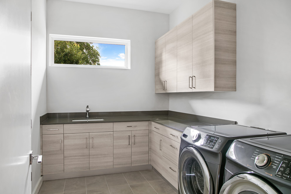 Bild på en mellanstor funkis grå l-formad grått tvättstuga enbart för tvätt, med en undermonterad diskho, släta luckor, bänkskiva i kvarts, en tvättmaskin och torktumlare bredvid varandra, grå väggar, beiget golv och grå skåp