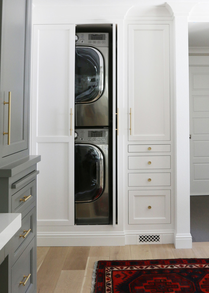 Diseño de armario lavadero pequeño con puertas de armario blancas, paredes blancas, suelo de madera clara y lavadora y secadora escondidas