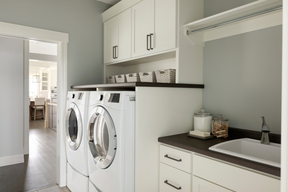 Modelo de cuarto de lavado lineal clásico renovado con fregadero encastrado, armarios estilo shaker, puertas de armario blancas, encimera de madera, paredes grises, lavadora y secadora juntas y encimeras marrones