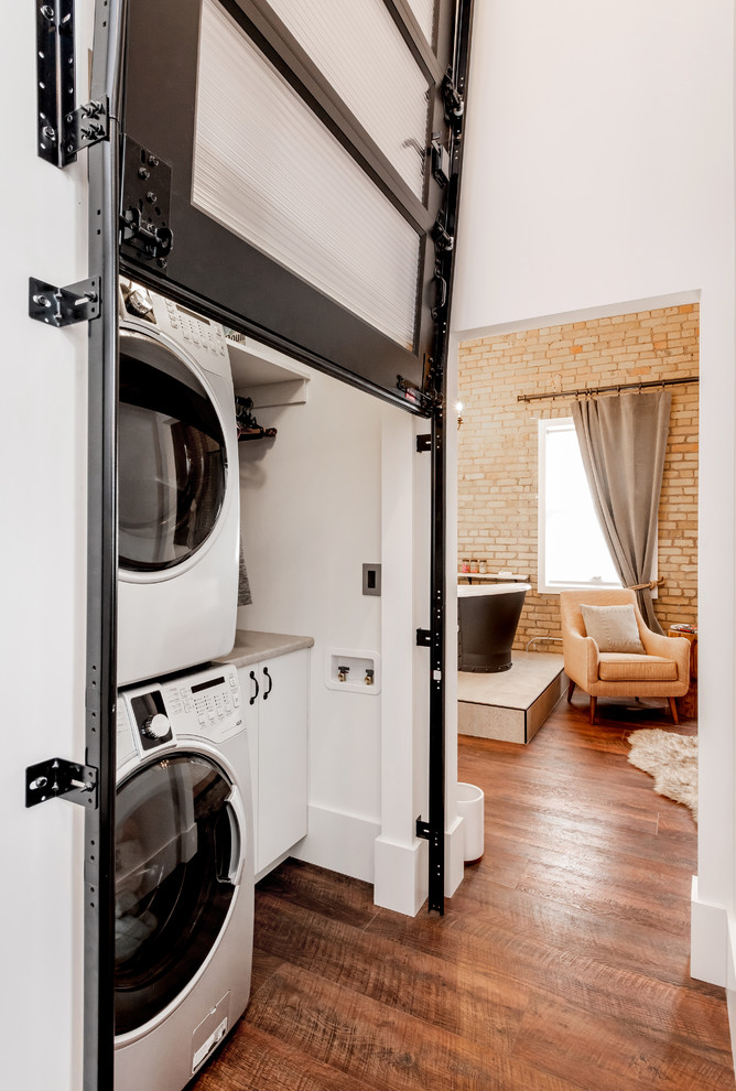 Ejemplo de armario lavadero lineal urbano pequeño con puertas de armario blancas, encimera de laminado, suelo laminado, lavadora y secadora apiladas, encimeras grises, paredes blancas y suelo marrón