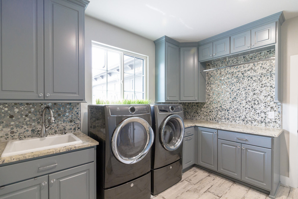 Lantlig inredning av ett grovkök, med blå skåp, granitbänkskiva, grå väggar, klinkergolv i porslin, en tvättmaskin och torktumlare bredvid varandra och grått golv