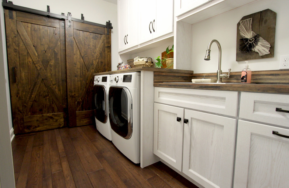 Rustik inredning av en mellanstor linjär tvättstuga enbart för tvätt, med en nedsänkt diskho, luckor med infälld panel, vita skåp, laminatbänkskiva, mörkt trägolv, en tvättmaskin och torktumlare bredvid varandra, brunt golv och vita väggar