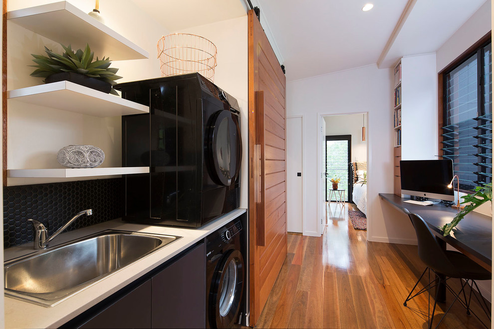 Bild på en liten funkis linjär liten tvättstuga, med en nedsänkt diskho, bänkskiva i kvarts, vita väggar, mellanmörkt trägolv, en tvättpelare, flerfärgat golv och svarta skåp