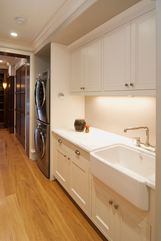 Small traditional single-wall utility room in San Francisco with a belfast sink, recessed-panel cabinets, white cabinets, marble worktops, beige walls, light hardwood flooring and a stacked washer and dryer.