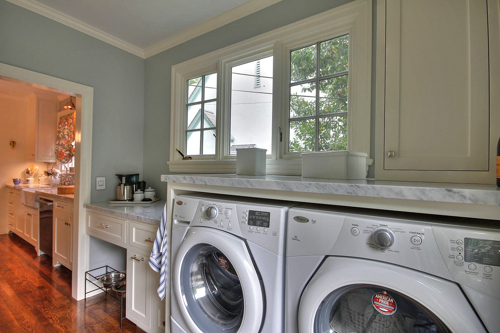 Modelo de lavadero multiusos clásico de tamaño medio con armarios estilo shaker, encimera de mármol, lavadora y secadora juntas, suelo de madera oscura, encimeras grises y paredes grises