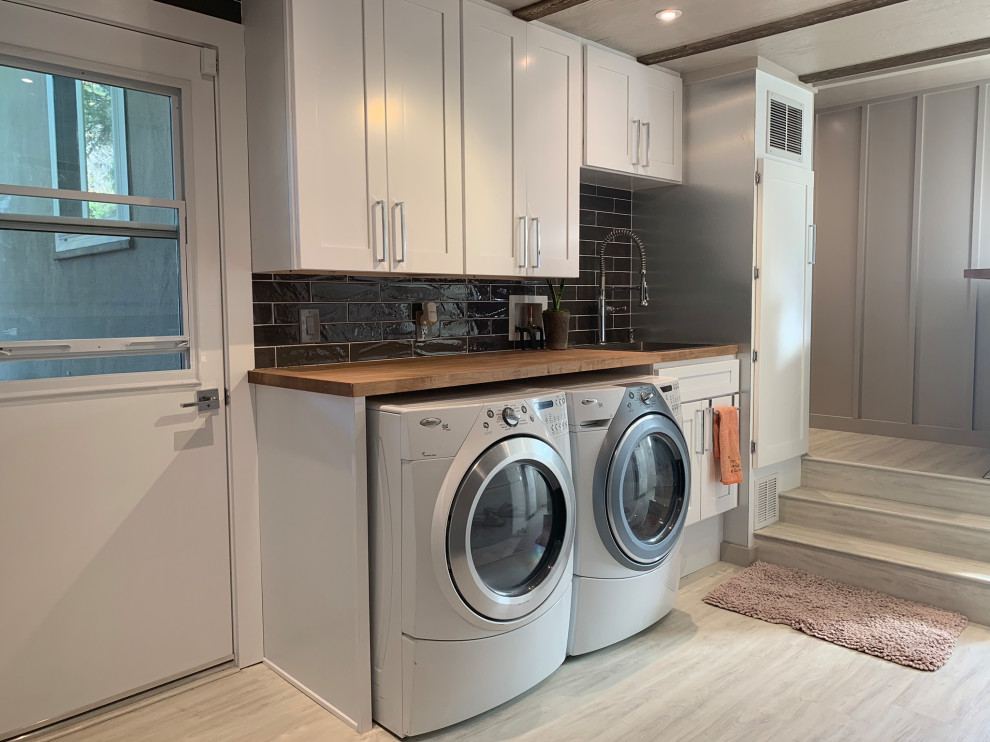 サンフランシスコにある低価格の中くらいなトラディショナルスタイルのおしゃれな家事室 (ll型、ドロップインシンク、シェーカースタイル扉のキャビネット、白いキャビネット、木材カウンター、グレーのキッチンパネル、セラミックタイルのキッチンパネル、グレーの壁、クッションフロア、左右配置の洗濯機・乾燥機、白い床、茶色いキッチンカウンター、表し梁、パネル壁) の写真