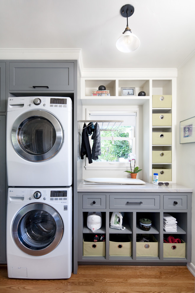 Modelo de lavadero lineal tradicional renovado grande con armarios estilo shaker, puertas de armario grises, paredes blancas, suelo de madera en tonos medios, lavadora y secadora apiladas y encimeras grises