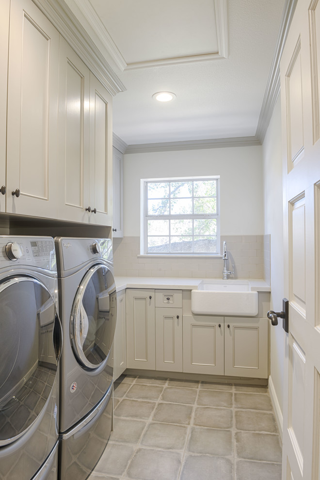 Klassisk inredning av en liten vita l-formad vitt tvättstuga enbart för tvätt, med en rustik diskho, luckor med profilerade fronter, beige skåp, vita väggar, klinkergolv i keramik, en tvättmaskin och torktumlare bredvid varandra och grått golv