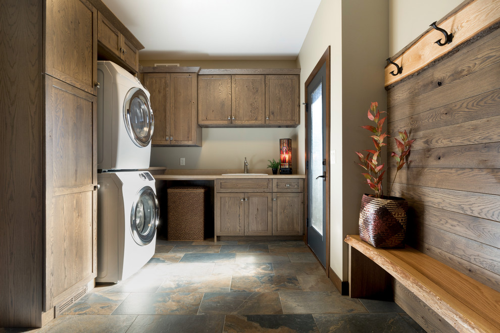 Urige Waschküche mit Einbauwaschbecken, Schrankfronten im Shaker-Stil, dunklen Holzschränken, beiger Wandfarbe, Waschmaschine und Trockner gestapelt und buntem Boden in Sonstige